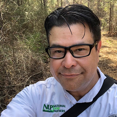 Close-up selfie of a man with dark hair and glasses, outdoors on a wooded path, wearing a white polo with a green National Partners logo and a black shoulder strap across his chest.