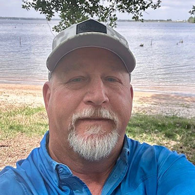 Portrait of a middle-aged man with a gray goatee, wearing a light gray cap and blue shirt, outdoors by a lake with a grassy shore and trees overhead.