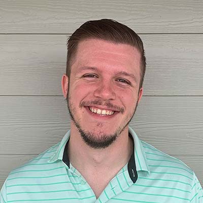 Smiling man with short hair and light beard wearing a mint green striped polo shirt, standing against a gray wooden wall.